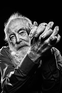 Uzun gri sakallı yaşlı bir adam ellerini kilitli tutar. Karanlık bir arka planda stüdyo resmi. Yaşlılık.