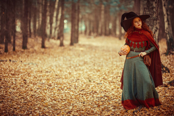 Halloween. Charming young girl witch with long red hair stands in a deep forest with a pumpkin. Fairy tales. Copy space.