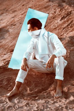 Beyaz elbiseli yakışıklı genç adam çölde ayna ile poz veriyor. Erkek modası.