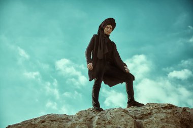 Maceralar, özgürlük ruhu. Cesur genç adam siyah elbiseler ve siyah türbanla tepenin üstünde bulutlu bir gökyüzüne karşı poz veriyor.. 