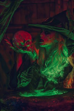 Cadılar Bayramı. Korkutucu yaşlı bir cadı eski ahşap bir evde kafatası tutuyor. Sihirli yeşil ve kırmızı ışık.