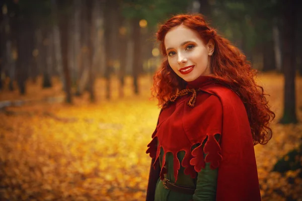 Beautiful red haired girl in a traditional green celtic dress and a red cloak is in forest. Historical reconstruction of ancient Celtic times.