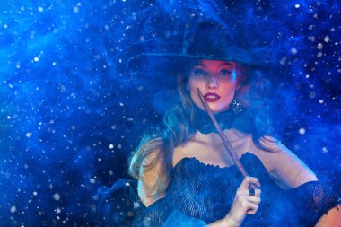 Cadılar Bayramı. Büyülü bir değnekle poz veren cadı şapkalı ve elbiseli muhteşem bir genç kadının portresi. Cadılar Bayramı partisi, makyaj ve kostüm..