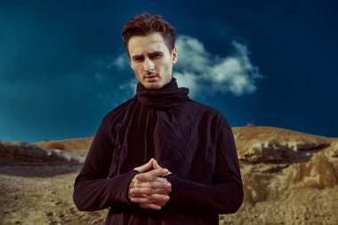 Çöl. Çölde duran siyah elbiseli yakışıklı bir esmer adam portresi. Erkek güzelliği, moda.