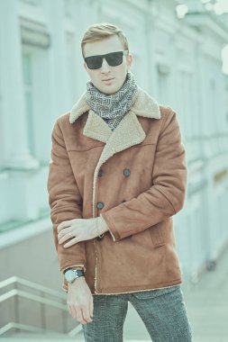 Erkek tarzı. Kışlık ceketli ve atkılı yakışıklı genç adam şehrin sokaklarında duruyor. Moda, giysi ve aksesuarlar..