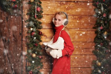 Kışlık elbiseli mutlu kız çocuk kaymaya gidiyor. Noel çelenkleri ve oyuncaklarıyla süslenmiş ahşap bir evin yanında kar yağışının altında duruyor. Kış aktiviteleri. 