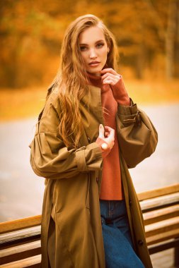 Sonbahar havası. Mevsimlik moda. Sonbahar kıyafetleri içinde güzel bir kız parkta bir bankın yanında duruyor, yaprak düşüyor.. 