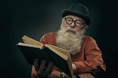 Melon şapkalı ve gözlüklü uzun gri sakallı bilge yaşlı bir adam eski bir kitap okuyor. Siyah arkaplan. 