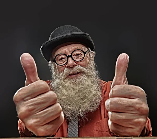 Old age. Portrait of a positive old-fashioned old man with a white beard in a bowler hat and round glasses showing thumbs and smiling. Black background.