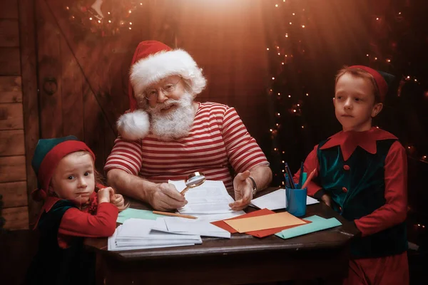 Santa Claus and little elves are sorting mail, reading letters from children. House, workshop of Santa Claus.