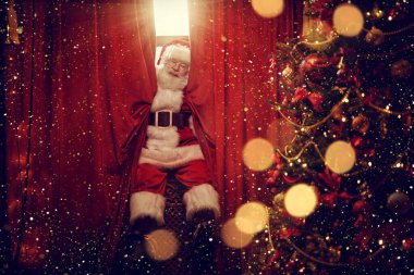Neşeli Noel Baba pencereden gizlice eve girer, pencere perdesine oturur ve perdelerin arkasından dışarı bakar. Noel ve Yeni Yıl kavramı.
