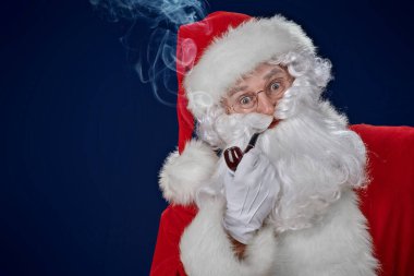 Noel ve Yeni Yıl. Pipo içen komik bir Noel Baba portresi ve şaşırtıcı. Stüdyo portresi koyu mavi arka planda. Boşluğu kopyala.