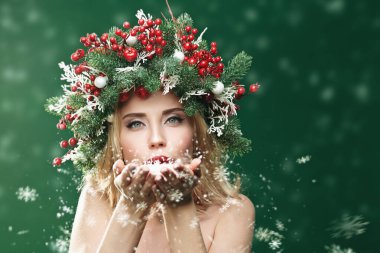 Çekici Noel kızı avuçlarından kar üflüyor. Noel atmosferi, ışıklar ve kar yağışı. Noel topları, böğürtlenler ve kar taneleriyle süslenmiş bir çam başlığı. Boşluğu kopyala.