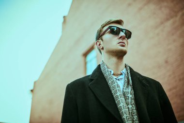 Şehir erkeğinin tarzı. Siyah güneş gözlüklü ve zarif siyah ceketli yakışıklı adam büyük şehrin grunge tuğlalı evlerinin arka planında duruyor.. 