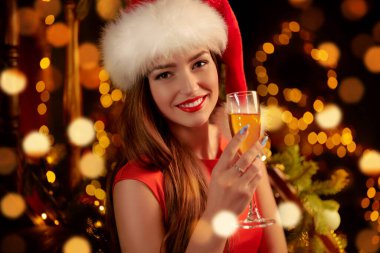 Yeni yıl ve Noel selamları. Kırmızı elbiseli ve Noel Baba şapkalı güzel kız bir bardak şampanya ve gülümseme tutuyor.. 