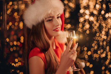 Yeni yıl ve Noel selamları. Kırmızı elbiseli ve Noel Baba şapkalı güzel kız bir bardak şampanya ve gülümseme tutuyor.. 