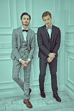 Men's style. Full length portrait of two elegant young men in classic three-piece suits standing in white luxury apartment. clipart