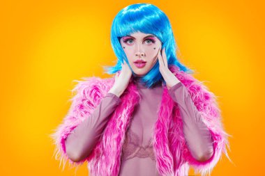 Parlak pembe makyajlı ve sarı arka planda pembe kürk giyen mavi peruklu çekici bir parti kızı portresi. Makyaj, makyaj, saç stili. Moda kızı. Boşluğu kopyala.