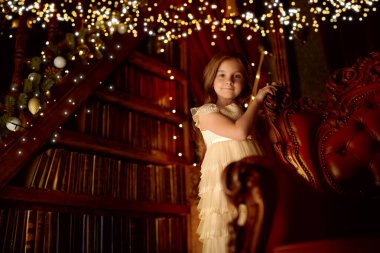 Sevimli küçük kız, altın ışıklarla dolu bir Noel atmosferinde eski bir koltuğun yanında duruyor. Noel Arifesi.