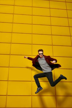 Palto ve güneş gözlüklü şık genç adam sarı sanayi duvarının oradaki bir sokakta koşuyor. Modern şehir yaşam tarzı. Erkek modası. 