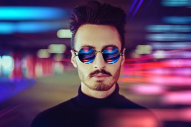 Siyah kazak ve yuvarlak güneş gözlüğü takan genç bir adamın portresi şehir ışıklarının arka planına karşıdır..