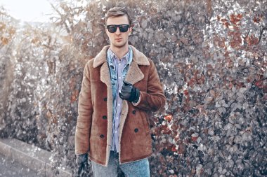 Koyun derisi ceketli yakışıklı genç adam kış parkında karın altında yürüyor. Erkek modası. 