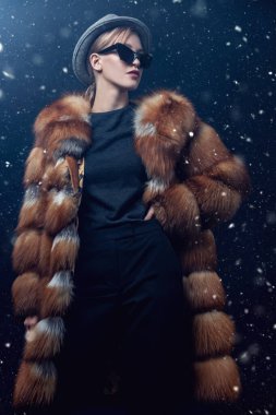 Lüks bir tilki kürkünün içinde, koyu mavi bir arka planda kar altında poz veren manken. Kış kürk manto modası.