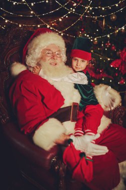 Nazik Noel Baba ve elfi evdeki koltukta dinleniyorlar. Noel ve Yeni Yıl masalları.