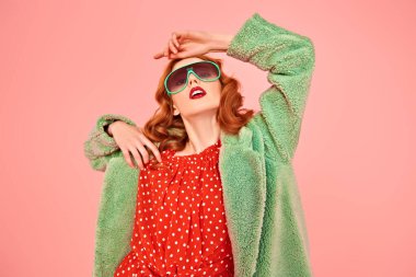 Kırmızı elbiseli, yeşil kürklü ve güneş gözlüklü şık bir genç kadının portresi. Pembe arka planda moda çekimi.