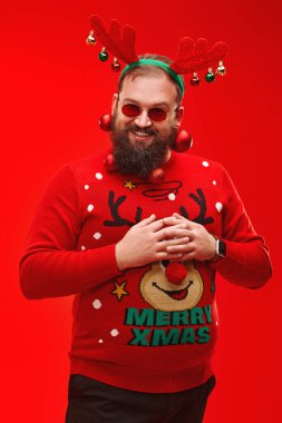 Kameraya gülümseyen Noel toplarıyla süslenmiş sakallı komik bir hippi portresi. Modern hippi Noel Baba. Kırmızı arkaplan.