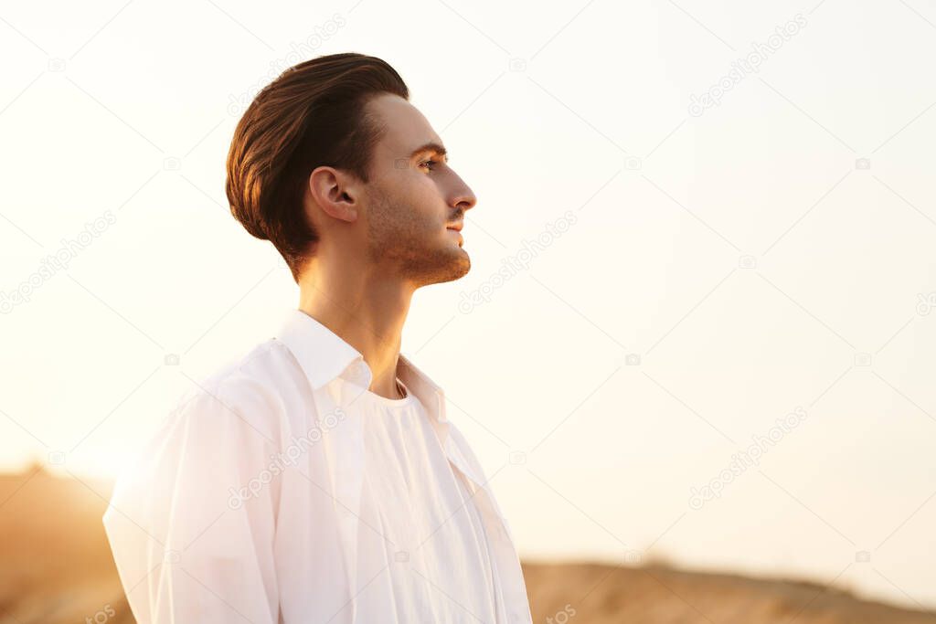 Portrait of a romantic young man in white clothes standing in a desert and looking at sunset. 