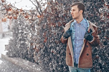 Koyun derisi ceketli yakışıklı genç adam kış parkında karın altında yürüyor. Erkek modası. 