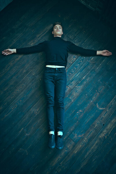 Красивый молодой человек в черной одежде лежит на черном деревянном полу, руки раскинуты по сторонам. Религиозное понятие.