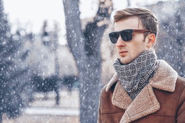 Güneş gözlüklü zarif bir adamın kış portresi ve kış parkının arka planında bir ceket. Kar yağışıweather condition. 