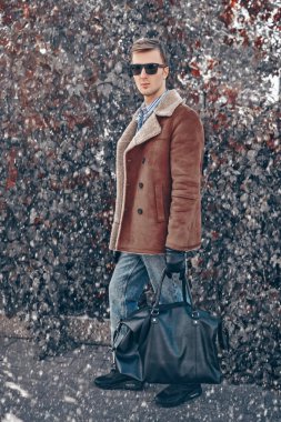 Kışlık ceketli yakışıklı genç adam ve siyah deri eldivenli siyah deri çantalı kış parkında poz veriyor. Erkek modası. Giysiler ve aksesuarlar. 