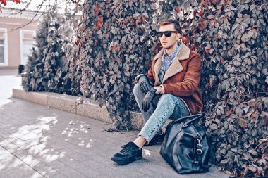 Sonbahar, şehir ve insanlar. Sonbahar ceketli ve güneş gözlüklü yakışıklı genç bir adam bir şehir caddesinde oturuyor. Erkek modası. Giysiler ve aksesuarlar. 