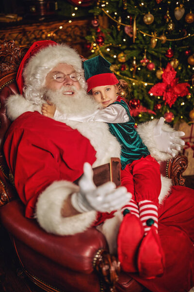 Новогодние и рождественские сказки. Добрый старый Санта Клаус и его эльф отдыхают в кресле дома. 