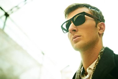 Şehir erkeğinin tarzı. Siyah güneş gözlüklü yakışıklı bir adamın yakın plan portresi gökyüzüne ve şehir arka planına karşı.. 