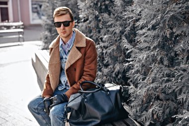 Kış, şehir ve insanlar. Kışlık montlu yakışıklı genç adam bir şehir caddesinde bir bankta oturuyor. Erkek modası. Giysiler ve aksesuarlar. 