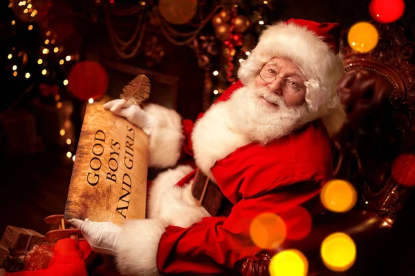 Happy Santa Claus bagging Christmas presents, checking out a list of wishes. He sits in an armchair in a beautiful Christmas interior. Christmas and New Year.