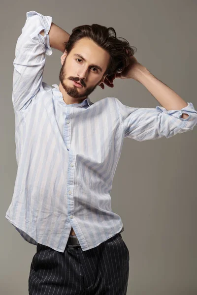 Men\'s beauty and hairstyle. Handsome brunet man posing at studio in light shirt and trousers. Men\'s fashion.