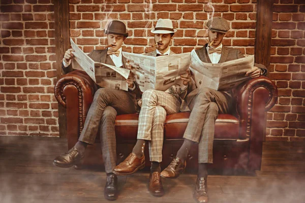 Three handsome men in elegant suits sit on a leather sofa, smoke cigars and read newspapers. Newspaper editorial office. Retro style. Men\'s fashion.