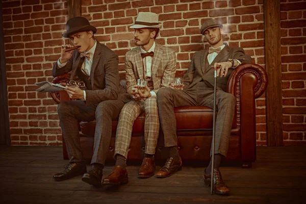 Three handsome men in elegant suits spends time in the men's club with cigars and whiskey. Luxury lifestyle. Men's beauty, fashion.