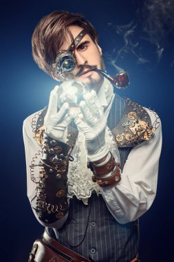 Hayal dünyası. Tek gözlü kristal enerji topuna bakan yakışıklı bir steampunk adamın portresi. Koyu mavi arkaplan.  