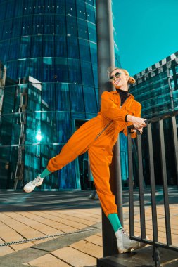 Parlak gençlik tarzı. Parlak turuncu tulumlu ve yuvarlak güneş gözlüklü modern moda kız gökdelenlerin arka planında poz veriyor. Moda fotoğrafı..