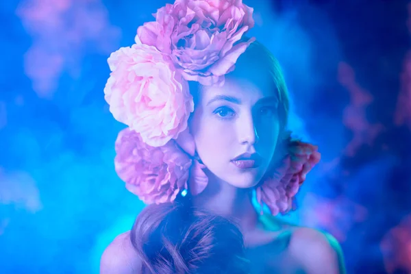 Fashion and flowers. Beautiful fashion model with peony flowers in her hair on a blue background with light haze. Make-up and hairstyle. Studio portrait.