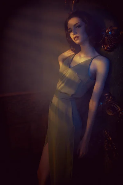 Portrait of a beautiful brunette woman in a light elegant dress posing in a dark vintage room lit by the rays of the setting sun. Beauty, fashion concept. Evening make-up and hairstyle.