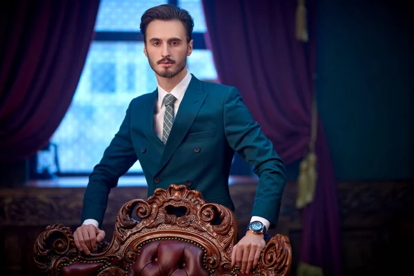 Luxury lifestyle. Portrait of a handsome young man in elegant classic suit standing by a vintage armchair in a luxury apartment. Men\'s beauty, fashion.