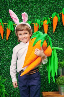Tavşan kulaklı mutlu küçük çocuk bahar bahçesinde havuç ve paskalya süslemeleriyle duruyor. Paskalya tatili.