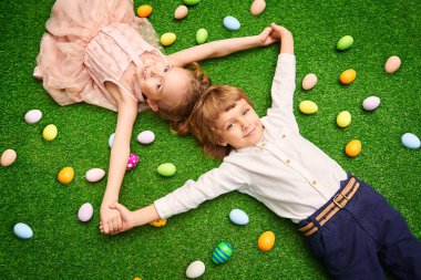 Sevimli gülücük oğlanı ve kız renkli yumurtalarla yeşil bir çimenlikte yatıyorlar. Paskalya tatili.